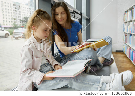 Mother and daughter reading books sitting on windowsill in library 123692966