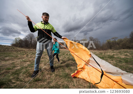 Setting up a vibrant tent on a cloudy afternoon in a serene field, as a child glimmers with excitement in the background during an outdoor adventure 123693080