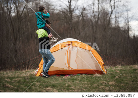 Enjoying a family camping adventure, a father carries his daughter on his shoulders while preparing a cozy orange tent in a serene natural setting during the cool afternoon 123693097