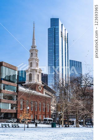 View of Park Street Church and surrounded buildings in Boston during winter with snow on the Boston Common ground and flock of birds flying against clear blue sky 123693965