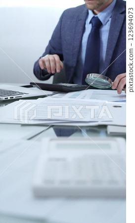 Businessman wearing blue suit is analyzing financial reports with magnifying glass and using calculator in a professional office setting. Low lighting, close-up vertical view. Business people, audit 123694073