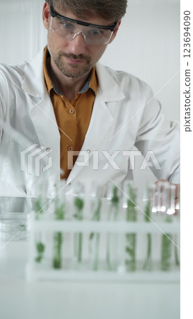 A male science researcher, wearing a lab coat and protective glasses, drips liquid from a pipette into a test tube containing a green plant. His face is focused, captured in a vertical portrait A male science researcher, wearing a lab coat and protective glasses, drips liquid from a pipette into a test tube containing a green plant. His face is focused, captured in a vertical portrait 123694090