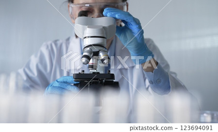 Woman scientist wearing lab coat, white protective gloves and glasses, is using microscope in modern laboratory setup, portrait view. Low key lighting photo. Science and Medicine 123694091