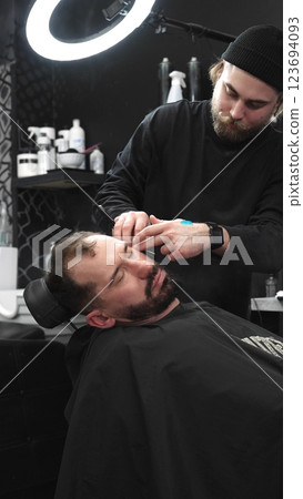 Professional barber carefully shaving client's beard with straight razor, applying gel and using hot towel in contemporary barbershop setting, vertical view 123694093