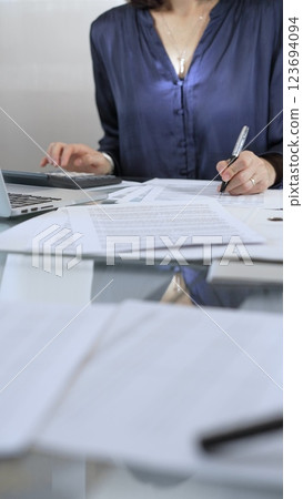 Businesswoman wearing blue dress is working with laptop, calculator and documents in a professional office setting. Low lighting, close-up vertical view. Business people, audit concept Businesswoman wearing blue dress is working with laptop, calculator and documents in a professional office setting. Low lighting, close-up vertical view. Business people, audit concept 123694094