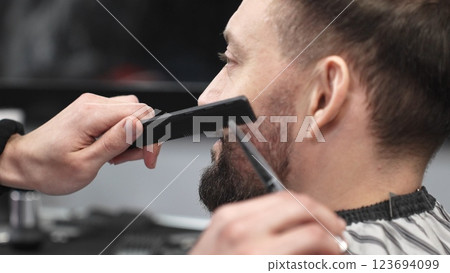 Skilled barber carefully trimming client's beard with precision scissors and sharp comb, working inside contemporary grooming salon 123694099