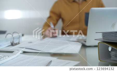Magnifying glass, calculator, pen and documents are lying on accountant's desk. Businessman or accountant, wearing yellow shirt, is working with documents on the background. Business people, audit Magnifying glass, calculator, pen and documents are lying on accountant's desk. Businessman or accountant, wearing yellow shirt, is working with documents on the background. Business people, audit 123694132