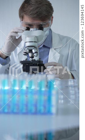 Male scientist researcher wearing lab coat, white protective gloves, medical mask and glasses, is using microscope in modern laboratory setup, portrait view. Low key lighting photo. Science 123694145