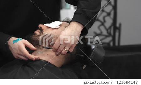 Professional barber carefully shaving client's beard with straight razor, applying gel and using hot towel for precise grooming in contemporary barbershop setting 123694148