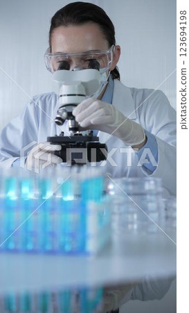 Woman scientist wearing lab coat, white protective gloves and glasses, is using microscope in modern laboratory setup, portrait view. Low key lighting photo. Science and Medicine concept 123694198