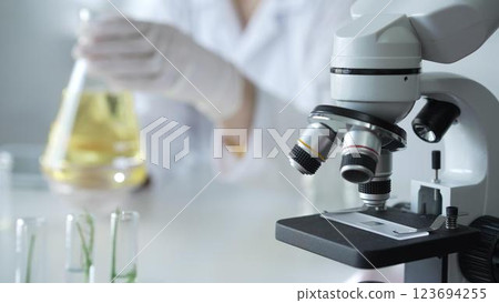 Microscope is near a scientist, wearing a lab coat and white protective gloves, and holding beaker with a yellow oily liquid in laboratory setting, close up. Medicine and science concept 123694255