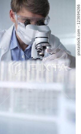 Man scientist wearing lab coat, white gloves, medical mask and protective glasses using microscope doing research in modern laboratory, portrait view. Low key lighting photo. Science and Medicine 123694266