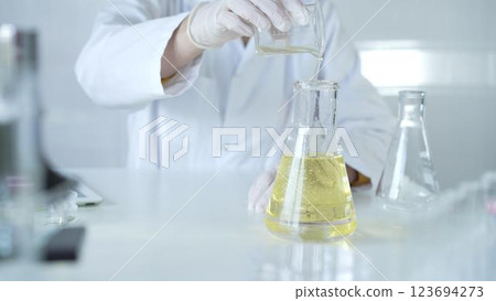 A scientist, wearing a lab coat and white protective gloves, is pouring a yellow oily liquid from one beaker to another in laboratory, close up. Medicine and science concept 123694273