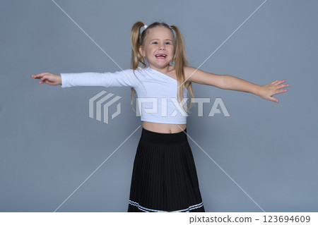 A cheerful young girl with blonde pigtails extends her arms wide, wearing a trendy white crop top and black pleated skirt. Her joyful expression conveys carefree childhood happiness. A cheerful young girl with blonde pigtails extends her arms wide, wearing a trendy white crop top and black pleated skirt. Her joyful expression conveys carefree childhood happiness. 123694609