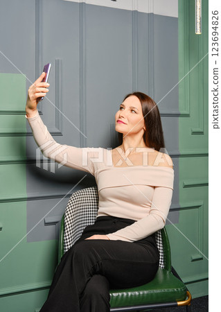 Middle-aged woman taking a selfie while sitting on a green chair in a stylish interior setting 123694826