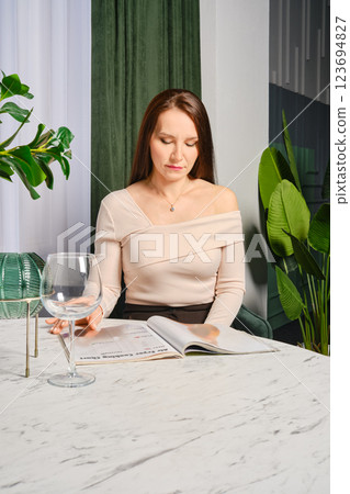 Middle-aged woman reading magazine while seated at a stylish table in a modern interior Middle-aged woman reading magazine while seated at a stylish table in a modern interior 123694827
