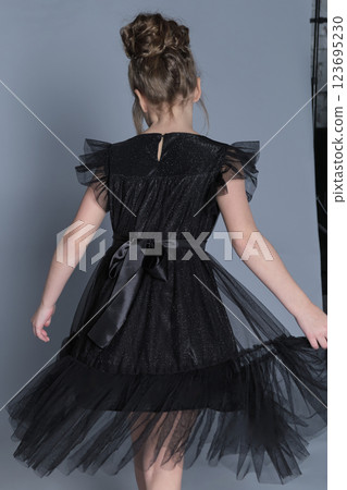 A young girl in a black tulle dress twirls gracefully, showing the elegant back details of her outfit. This image captures the beauty of movement in children's formal fashion, highlighting the dress's A young girl in a black tulle dress twirls gracefully, showing the elegant back details of her outfit. This image captures the beauty of movement in children's formal fashion, highlighting the dress's 123695230