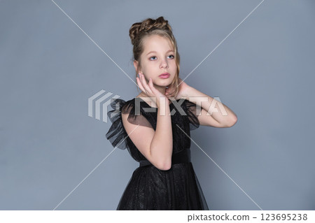 A girl in a formal black dress gently frames her face with her hands, embodying grace and beauty. The photo captures the essence of youthful elegance, emphasizing modern trends in children's fashion 123695238