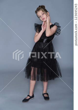 A girl stands gracefully in a black ruffled dress, lifting the fabric to reveal the delicate tulle layers. The photo emphasizes elegance and poise, highlighting modern trends in children's formal wear A girl stands gracefully in a black ruffled dress, lifting the fabric to reveal the delicate tulle layers. The photo emphasizes elegance and poise, highlighting modern trends in children's formal wear 123695243