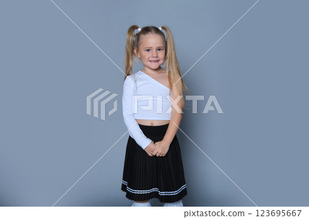 Wearing a white one-shoulder top and a black pleated skirt, a young girl with blonde pigtails grins mischievously. This image captures her playful energy and the innocence of childhood. 123695667