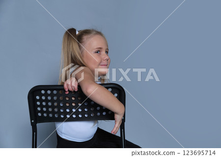 A playful 5-year-old girl is photographed in a studio, sitting on a black chair with a big, joyful expression. Dressed in a stylish one-shoulder top and black pleated skirt, she radiates energy and 123695714