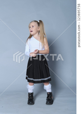 young girl showcases a chic look in a one-shoulder white top paired with a black pleated skirt and black boots. Her confident stance and playful expression bring out the modern trends in children's 123695738
