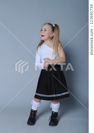 young girl with blonde pigtails is standing confidently against a grey background, wearing a stylish one-shoulder white top, a black pleated skirt, white lace socks, and black boots. Her pose is 123695739