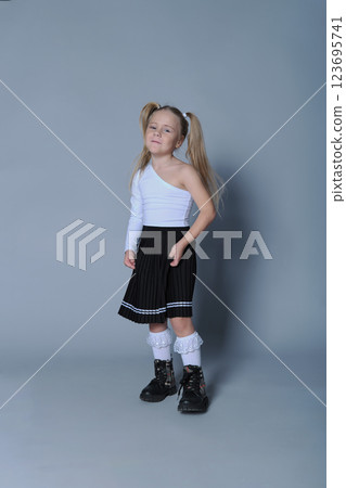 young girl with blonde pigtails is standing confidently against a grey background, wearing a stylish one-shoulder white top, a black pleated skirt, white lace socks, and black boots. Her pose is 123695741