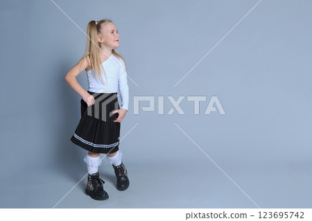cheerful young girl with blonde pigtails poses playfully against a plain grey background, dressed in a trendy one-shoulder white top, black pleated skirt, and lace-trimmed white socks paired with 123695742