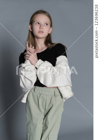A young girl with long blonde hair, dressed in a black top and light green pants, stands thoughtfully with her hands clasped. This image conveys a sense of contemplation and innocence, reflecting 123696238