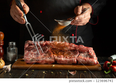 A chef meticulously seasons a large cut of meat using a strainer and a fork. Fresh herbs and spices are prominently present on the wooden board 123696741