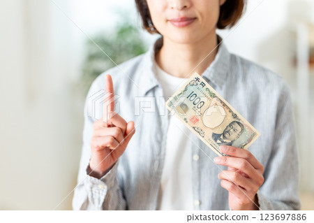 A middle-aged woman holding a 1,000 yen bill and pointing her index finger up 123697886