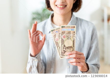 一名中年婦女手持兩張一萬日元鈔票,面帶微笑,畫著圓圈手勢 一名中年婦女手持兩張一萬日元鈔票,面帶微笑,畫著圓圈手勢 123698014