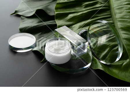 Cosmetic cream in a glass container rests on a large tropical leaf against a clean backdrop. 123700569