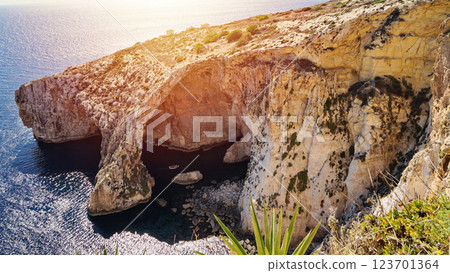 Stunning view of Blue Grotto in Malta, showcasing dramatic limestone cliffs, crystal-clear blue waters, and a small boat navigating the cave entrance under golden sunlight 123701364