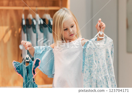 Child, fashion and shopping while choosing kids clothing in a shop, store or boutique with a cute girl holding dresses thinking or picking color choice. Little kid shopper or customer buying clothes Child, fashion and shopping while choosing kids clothing in a shop, store or boutique with a cute girl holding dresses thinking or picking color choice. Little kid shopper or customer buying clothes 123704171
