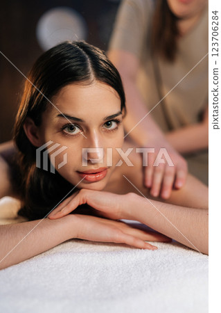 Vertical portrait of young woman relaxing with hand massage at beauty spa lying looking at camera. Professional masseur giving back massage to relaxed female client experiencing therapeutic wellness. Vertical portrait of young woman relaxing with hand massage at beauty spa lying looking at camera. Professional masseur giving back massage to relaxed female client experiencing therapeutic wellness. 123706284