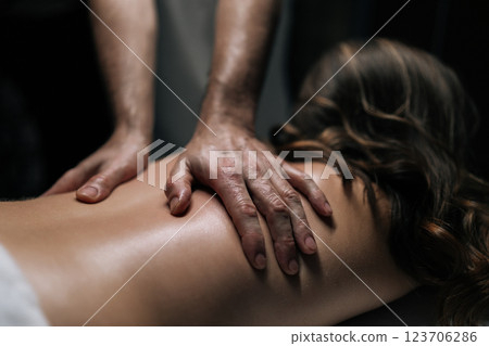 Close-up cropped shot of unrecognizable masseur performing deep tissue technique on female back, targeting muscle tension and promoting wellness during bodywork session in spa with dark interior. Close-up cropped shot of unrecognizable masseur performing deep tissue technique on female back, targeting muscle tension and promoting wellness during bodywork session in spa with dark interior. 123706286