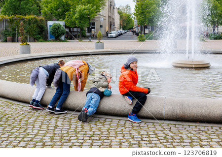 Group of children in colorful jackets gather around circular fountain in Potsdam, enjoying playful moment. Two lean over water, others observe lively city atmosphere, trees, and scenic views 123706819
