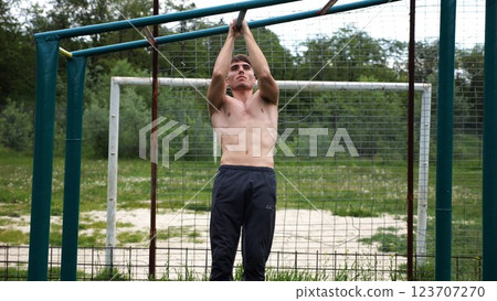 Young muscular sportsman doing pull ups at sports ground. Athletic guy exercising outdoor. Strong sporty man working out at summer nature. Concept of healthy and active lifestyle. Slow motion Young muscular sportsman doing pull ups at sports ground. Athletic guy exercising outdoor. Strong sporty man working out at summer nature. Concept of healthy and active lifestyle. Slow motion 123707270