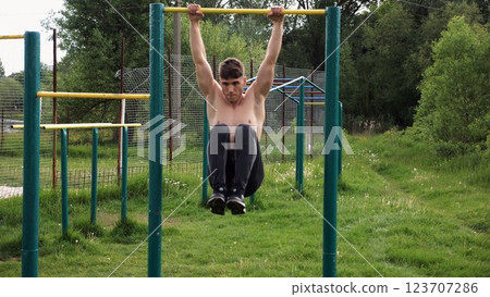 Strong guy training abdominal muscles on horizontal bar at sports ground. Young sportsman exercising on pull-up bar outdoor. Sporty athlete working out at nature. Concept of sportive lifestyle Strong guy training abdominal muscles on horizontal bar at sports ground. Young sportsman exercising on pull-up bar outdoor. Sporty athlete working out at nature. Concept of sportive lifestyle 123707286