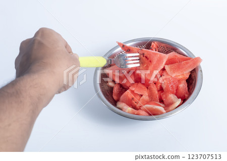 Watermelon slices in a stainless steel bowl with a white background, held by a hand and picked with a fork. 123707513