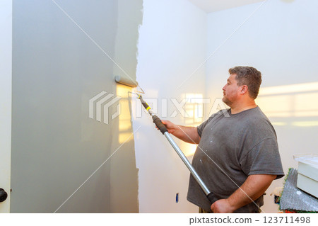 Man applies gray paint to white wall using roller in well lit room. 123711498