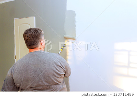 Contractor applies gray paint to wall using roller in well lit room, showcasing home improvement project. 123711499