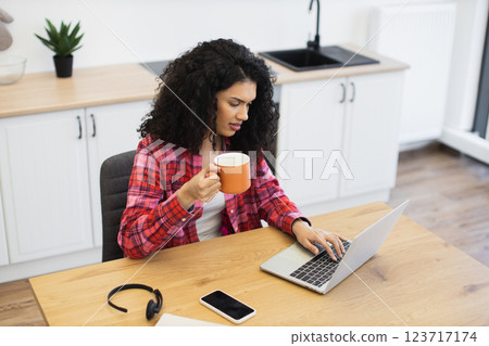 African woman early 30s working on laptop, drinking coffee, seated in kitchen workspace 123717174