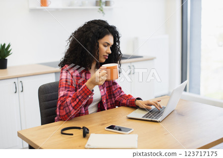 African woman early 30s working on laptop, drinking coffee, seated in kitchen workspace African woman early 30s working on laptop, drinking coffee, seated in kitchen workspace 123717175
