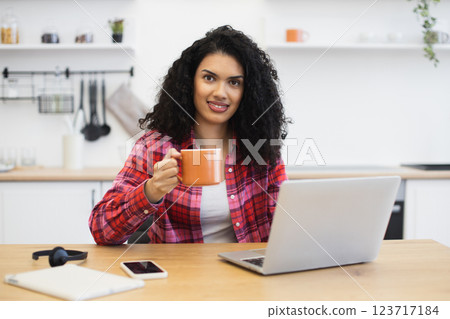 Smiling African woman with curly hair, wearing red shirt, working on laptop, holding coffee mug at home kitchen. Scene conveys comfort, productivity, and warmth. 123717184