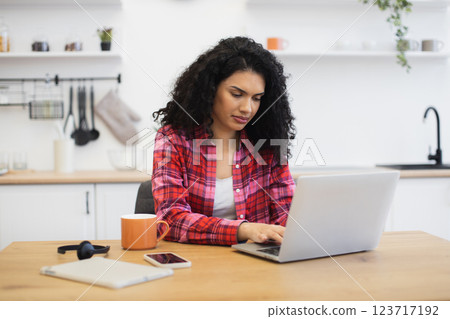 Young African woman sitting at kitchen table using laptop for remote work or study. Casual home environment with focus on technology and productivity. 123717192