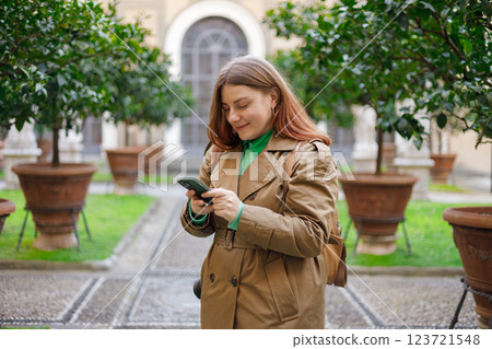 Picture of pretty young woman staying on the street holding phone in hands. 30s tourist in dress walking on old city street checks her smartphone. Technology concept, Traveling Europe, Italy in spring 123721548