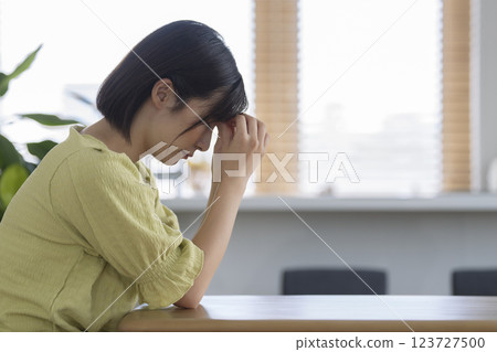 一位 30 多歲的女性在家中的餐桌旁抱著頭,側面 一位 30 多歲的女性在家中的餐桌旁抱著頭,側面 123727500